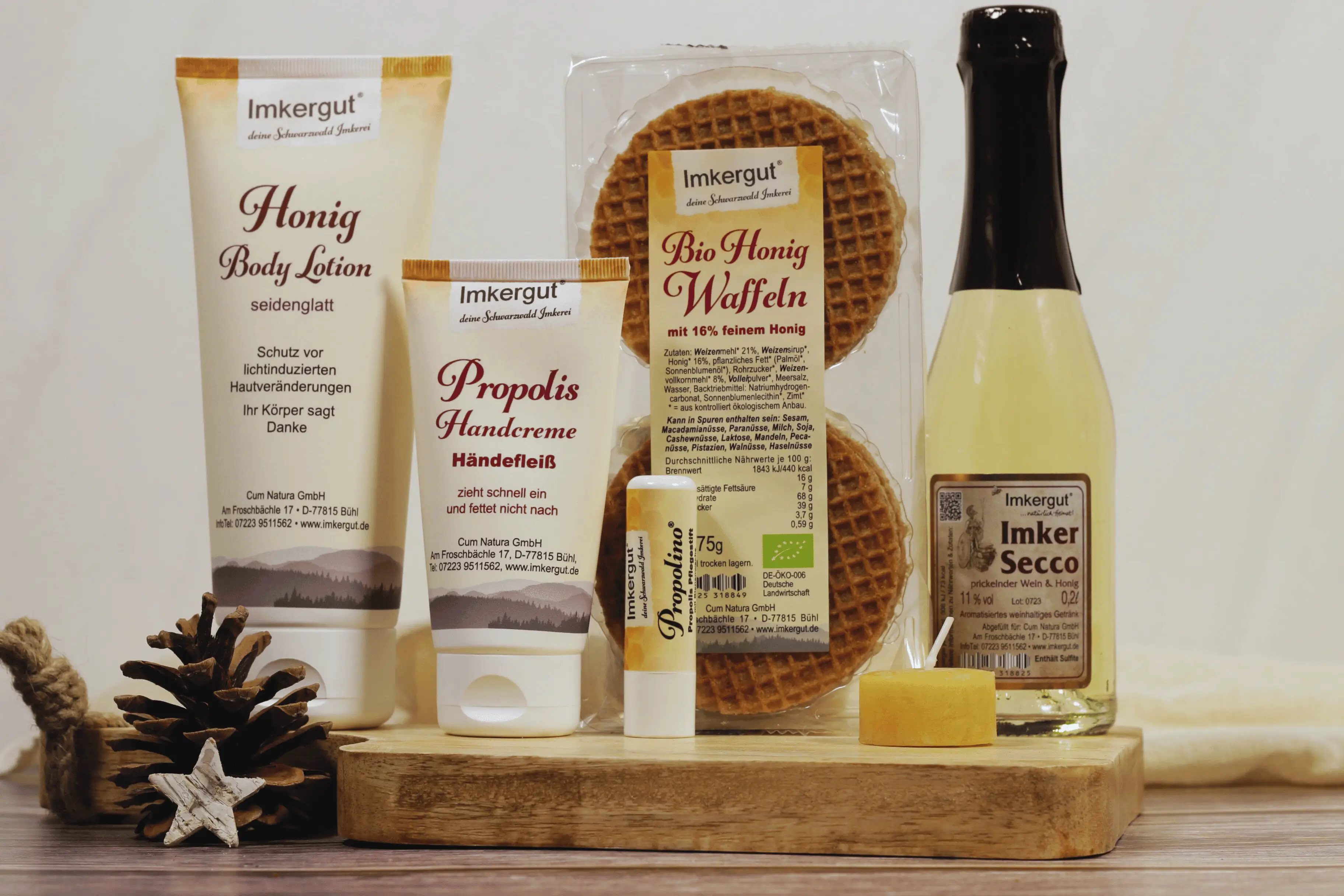 Eine Body Lotion, eine Handcreme, ein Lippenpflegestift, Honigwaffeln, ein Bienenwachsteelicht und ein Imker-Secco auf einem Holzbrett